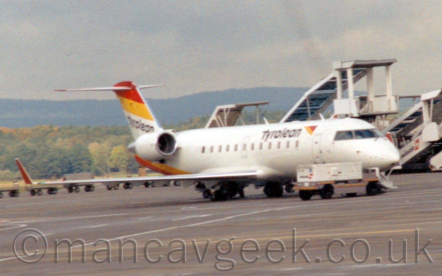 Side view of a twin engined jet airliner with the engines mounted on the sides of the rear fuselage, parked facing to the right. The plane is largely whit, with a yellow and orange triangle on the upper forward fuselage, next to black "Tyrolean" titles. Orange and yellow diagonal stripes on the lower rear fuselage, mirrored with orange and yellow diagonal stripes  over black "Tyrolean" titles. A white Ground Power Unit on a yellow trailer is parked in front of the nose of the plane. Grey concrete apron fills the foreground, with white mobile airstairs with large awnings parked on the right of the background, with tees in the distance on the left. Trees on a hill in the far distance are slowly vanishing into the grim grey sky which fills the rest of the frame.