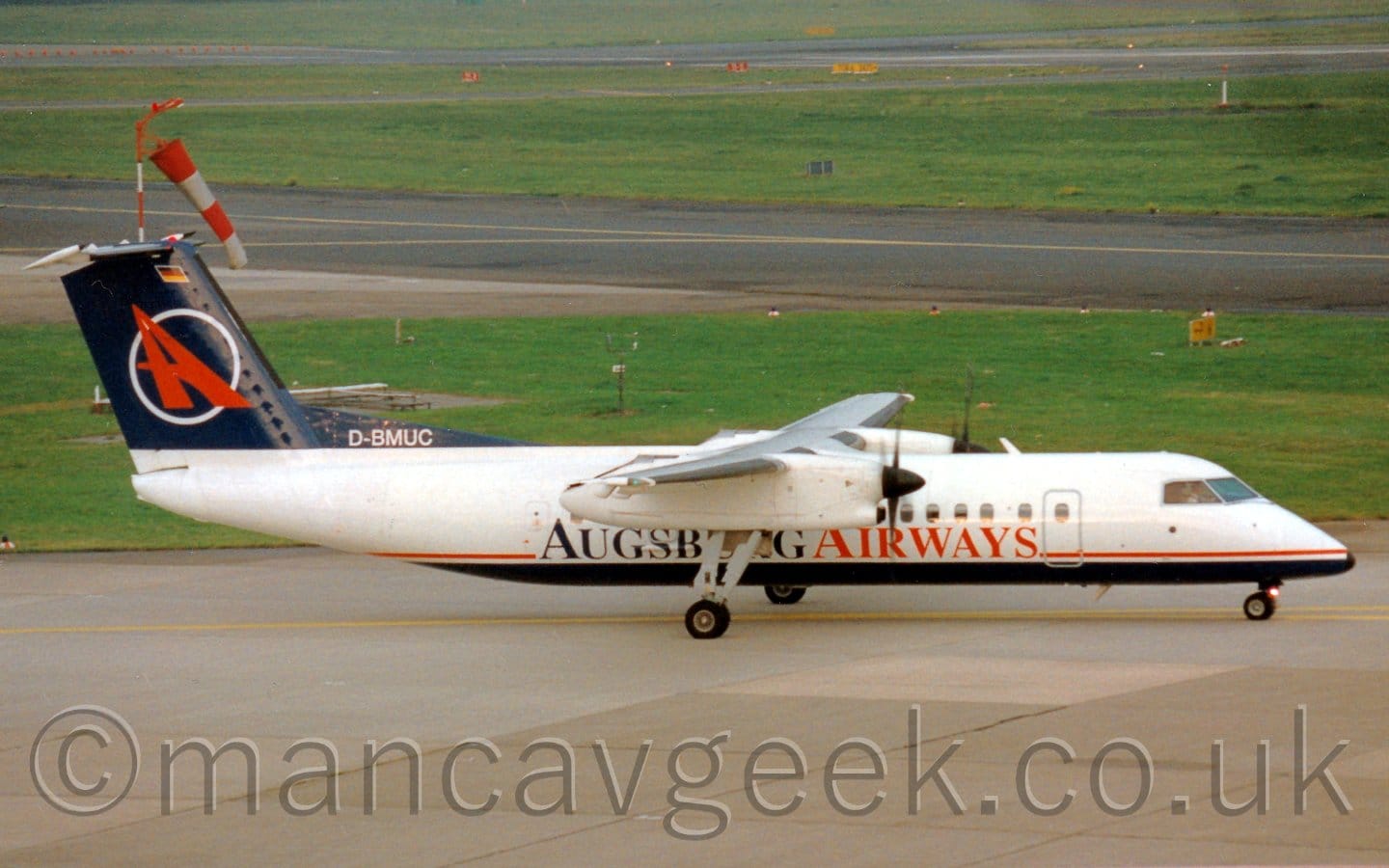 Side view of a high-winged, twin propellor-engined airliner taxiing from left to right. The plane is largely white, with a thin red stripe on the lower fuselage, a dark blue belly, and dark blue and red "Augsburg Airways" under the passenger cabin windows, in the middle of the body. The white registration "D-BMUC" is on a dark blue fillet at the bottom of the tail, which is also dark blue, and has the white outline of a circle containing a red upper-case "A", which leans to the 11 o'clock position. Grey concrete apron fills the foreground, with large grassed areas split by black taxiways and a runway in the background.