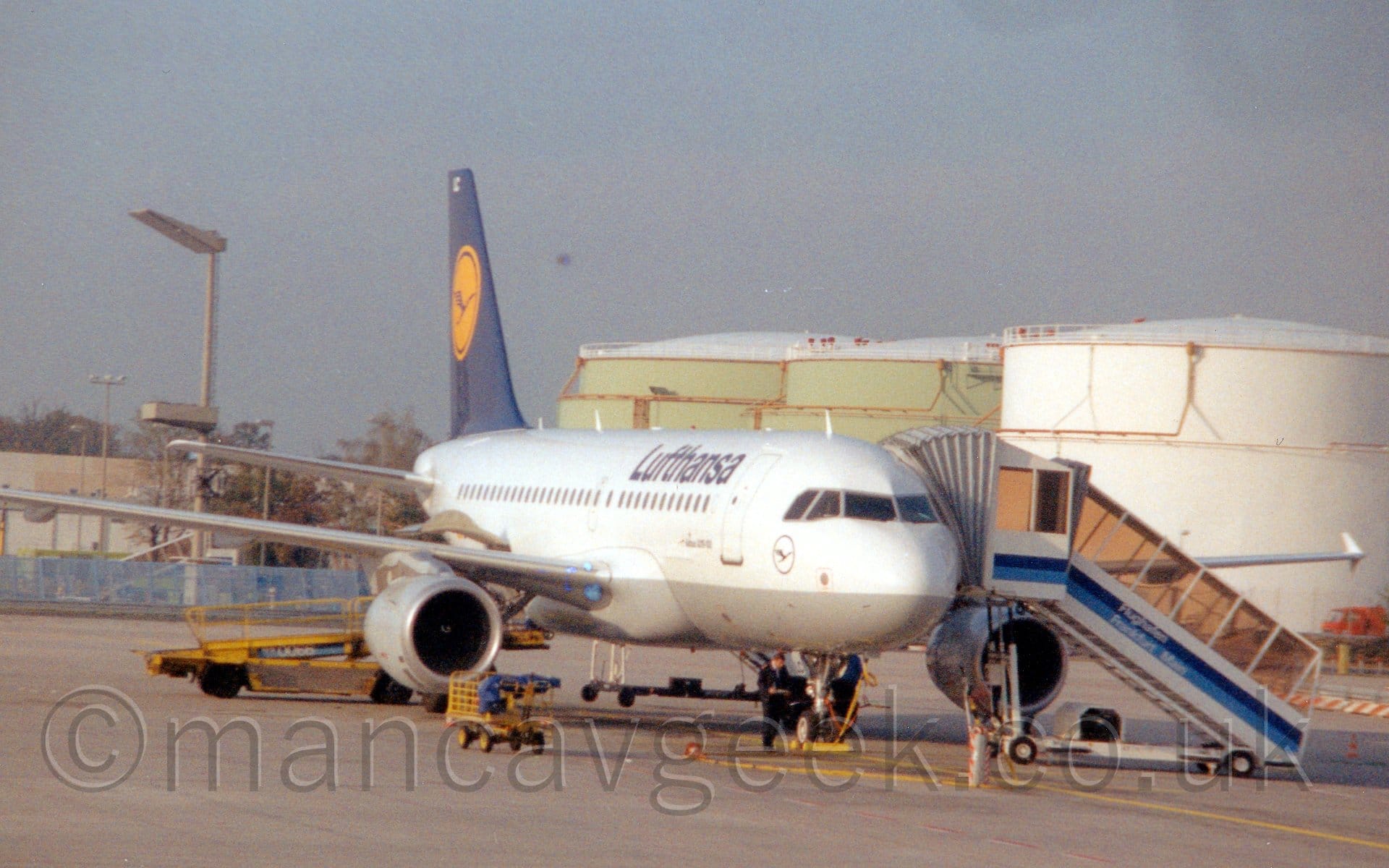 Side view of a twin engined jet airliner parked facing to the right and slightly towards the camera. The plane is mostly white, with a grey belly, with dark blue "Lufthansa" titles on the upper forwards fuselage. The black letters "LC" are on the open nose wheel doors. The dark blue tail has a yellow circle in the middle, containing a stylised blue flying bird. Yellow baggage carts and loading belts are scattered around the plane, with a white set of mobile airstairs with a blue stripe and "Flughafen Frankfurt Airport" titles on the side. Grey concrete apron fills the foreground, with large, circular green and white fuel tanks on the right of the frame, and tall lighting poles and trees on the left, under flat grey sky with patches of blue.