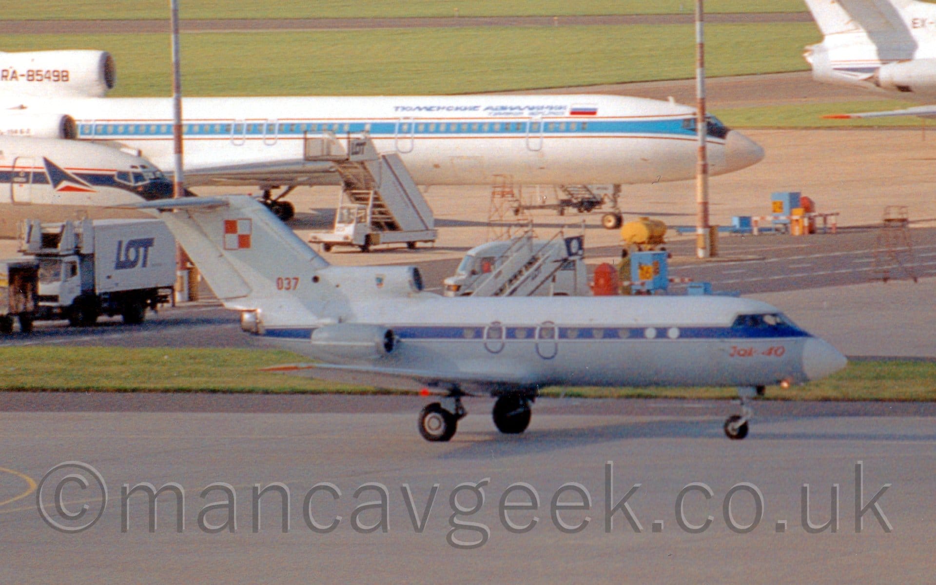 Side view of a small, 3 engined jet airliner in military colours taxiing from left to right. The plane is white on top with a thick blue stripe running along the body and a grey lower fuselage, with red "Yak-40" titles under the cockpit. The red serial "037" is on the bottom of the tail, with a 2*2 grid of red and white squares, outlined in red and white, at the top. The engine pods mounted on the sides of the rear fuselage are grey, while the centre engine intake between the top of the fuselage and the tail is white. Grey apron fills the foreground, with more apron filled with various white and blue jet airliners in the background, with a pair of red and white striped lighting poles stretching into the sky. Green grass lines runways, stretching out into the distance.