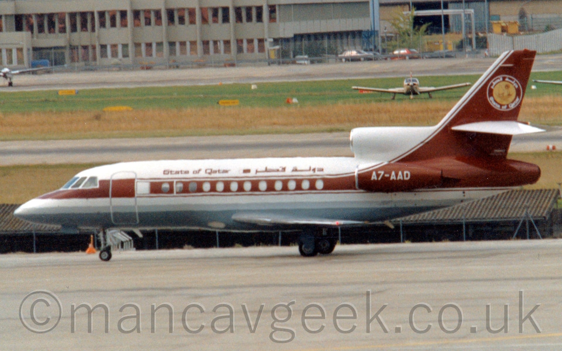 Side view of a 3 engined BizJet taxiing from right to left. The plane is largely white, with a maroon stripe running along the body and up into the tail, and a grey belly. The white registration "A7-AAD" is on the side of the maroon engine pods mounted on the rear fuselage. There are maroon "State of Qatar" titles on the upper fuselage, in English at the front and in an Arabic script towards the rear. A yellow circle with white "State of Qatar" titles in English at the bottom and in Arabic at the top is at the top of the tail. Grey concrete apron fills the foreground, with grass lining the sides of a black runway in the background. A single engined light aircraft is parked on the grass on the far side of the runway. A large grey building stretches across the frame in the background.