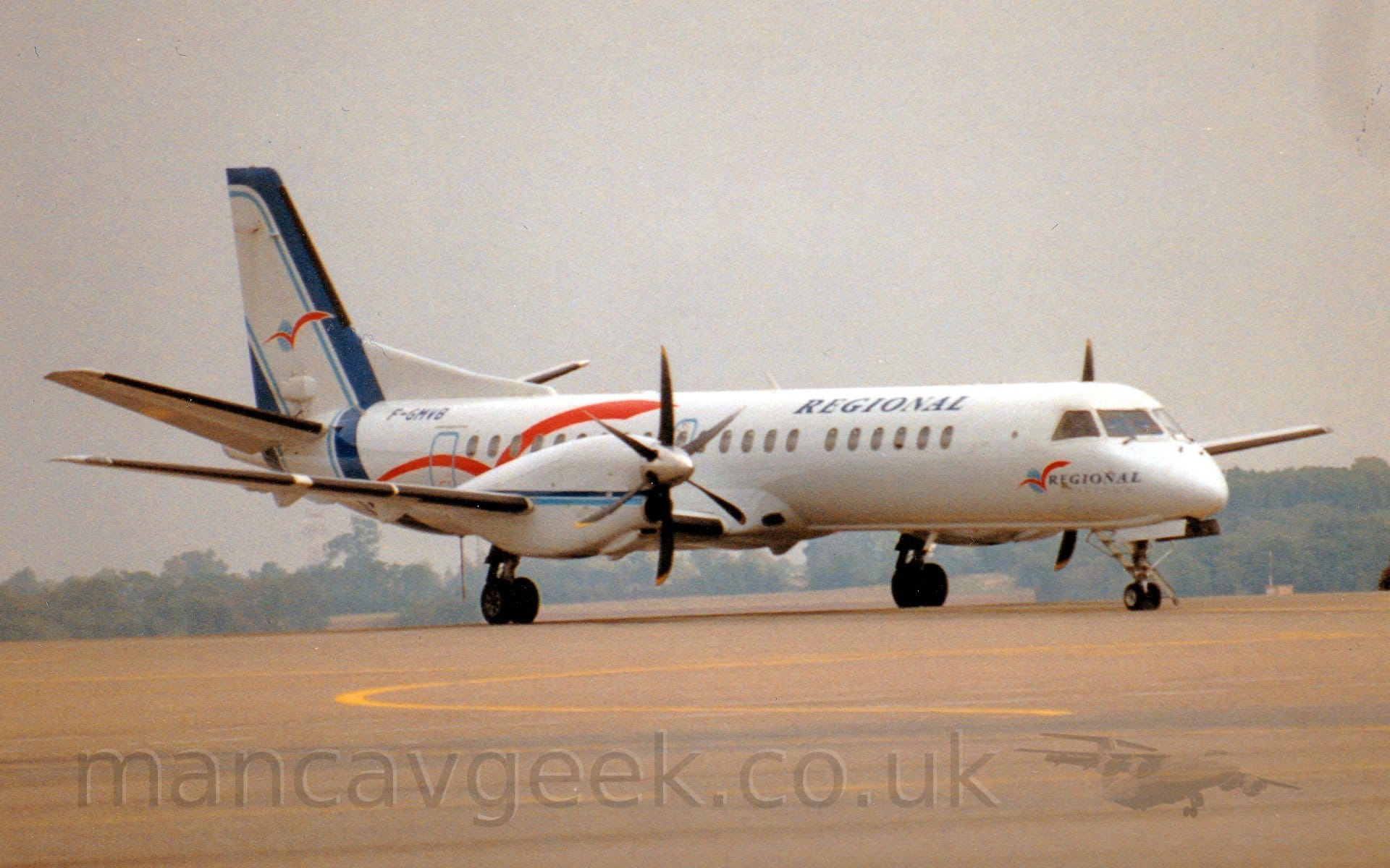 Side view of a twin propellor-engined airliner parked facing to the right. The plane is mostly white, with diagonal dark blue stripes with light blue piping along the leading and trailing edges of the tail, running down to loop under the rear fuselage. Overlaid with a stylised red flying bird over a blue ord. A larger version of the red bird logo adorns the rear fuselage, as well as a smaller version with small "Regional" titles under the cockpit. There are larger "Regional" titles on the upper forward fuselage, and the black registration "F-GMVB" on the upper rear. Thin dark and light blue stripes run along the length of the engine pods in front of the wings. Sandy-grey concrete apron marked with yellow lines for planes to follow fills the foreground, with trees in the distance, under grim, hazy sky.
