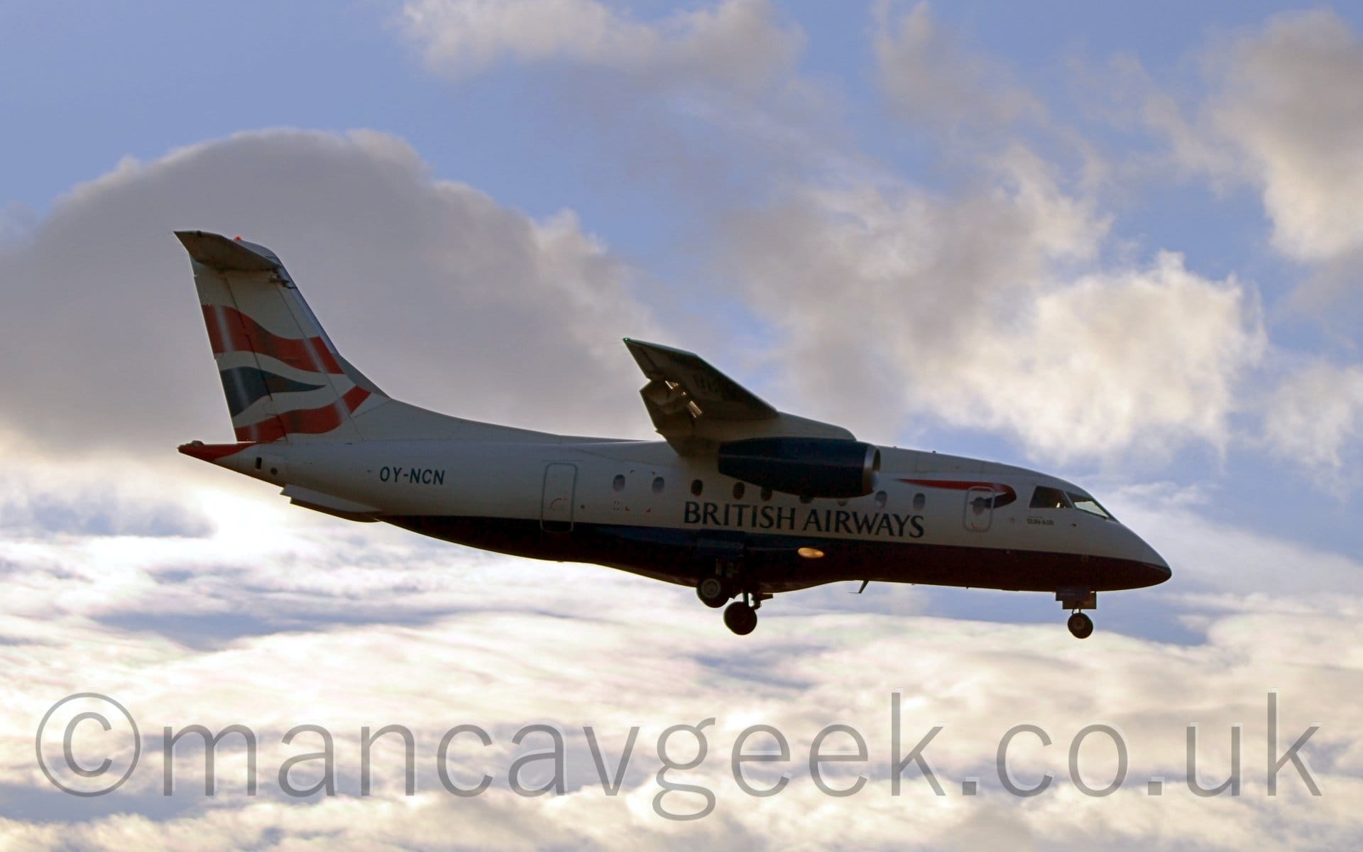 Side view of a high-winged, twin engined jet airliner flying from left to right at a low altitude, with undercarriage extended and flaps deployed from the rear of the wings, suggesting it is about to land. The plane is largely white, with a blue belly, and black "British Airways" titles on the lower centre fuselage. The engines slung under the wings are the same dark blue, and there is a twisted dark blue and red ribbon on the upper forward fuselage, behind the cockpit. The tail is white, with a pair of wavy red lines with a wavy dark blue triangle between them. Fluffy white clouds with patches of pale blue sky fill the rest of the frame.