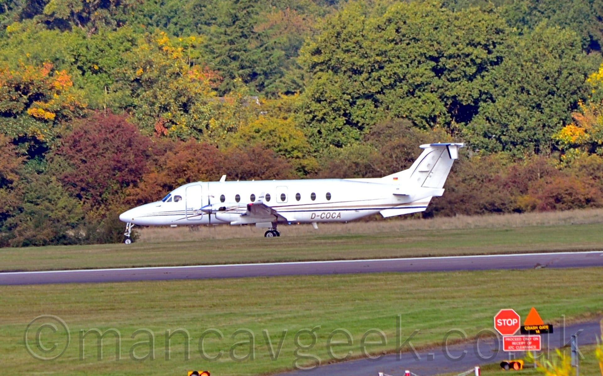 Slightly over-exposed side view of a twin propellor engined airliner taxiing from right to left. The plane is white, with a thin black and gold stripe running along the body, and the golden registration "D-COCA" on the lower rear fuselage. Green grass fills the foreground, lining both sides of a black runway. Trees fill the background, with a grey house barely visible through the foliage.