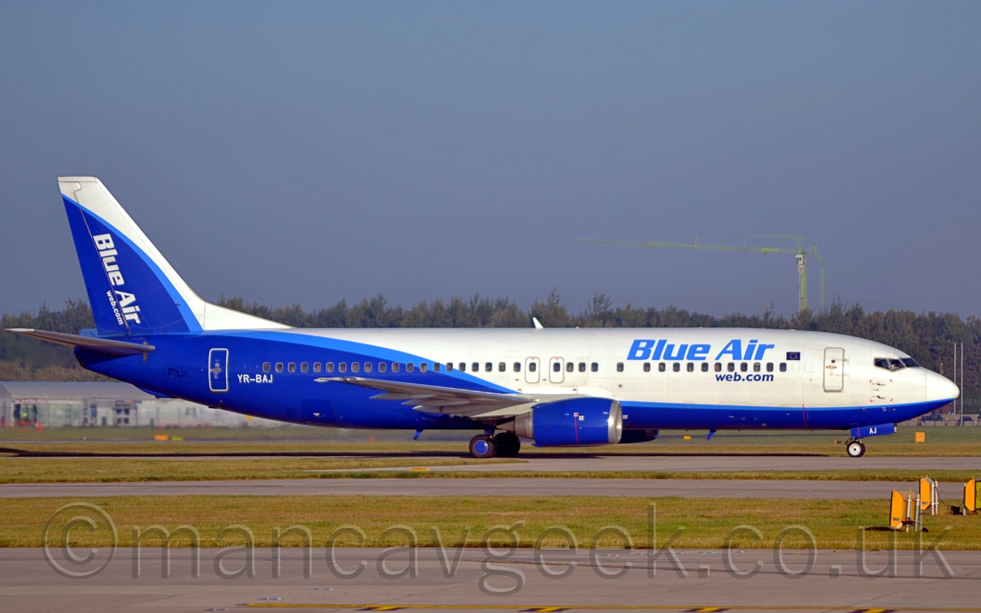 Side view of a twin engined jet airliner taxiing from left to right. The plane is mostly white, with a blue belly at the front, although the rear is mostly blue with a white roof, with a light blue stripe separating the 2 sections. There are blue "Blue Air" titles on the upper forward fuselage, above the passenger cabin windows, with smaller, darker "web.com" web address below the windows. The white registration "YR-BAJ" is on the lower rear fuselage., just ahead of the aft cabin door. The tail is mostly white, with a white leading edge, and white diagonal "Blue Air" titles and "Web.com" web address. A grey runway and taxiway, lined with green grass, fills the foreground, while trees stretch across the background, under blue-grey sky.