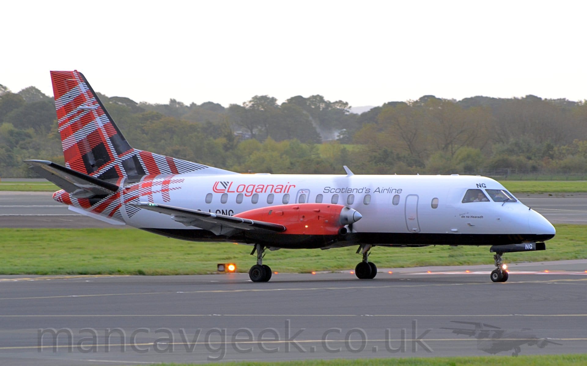 Side view of a twin propellor-engined airliner taxiing along a black taxiway from left to right. The plane is largely white, with a black belly, and the black letters "NG" above the cockpit windows, repeatedin white on the open nose-weheel doors. There are black "Scotlands Airline" titles on the upper forward fuselage, with larger, red "LoganAir" titles on the upper rear. The black registration "G-GNG" is on the lower rear fuselage, partially obscured by the wing. The rear fuselage and tail is covered in a red, black, and white tartan. The engine pods on the wings are a bright red. Black taxiway fills the foreground, lined by green grass. A black runway stretches across the background, with trees leading off into the distance, under bright but hazy sky.