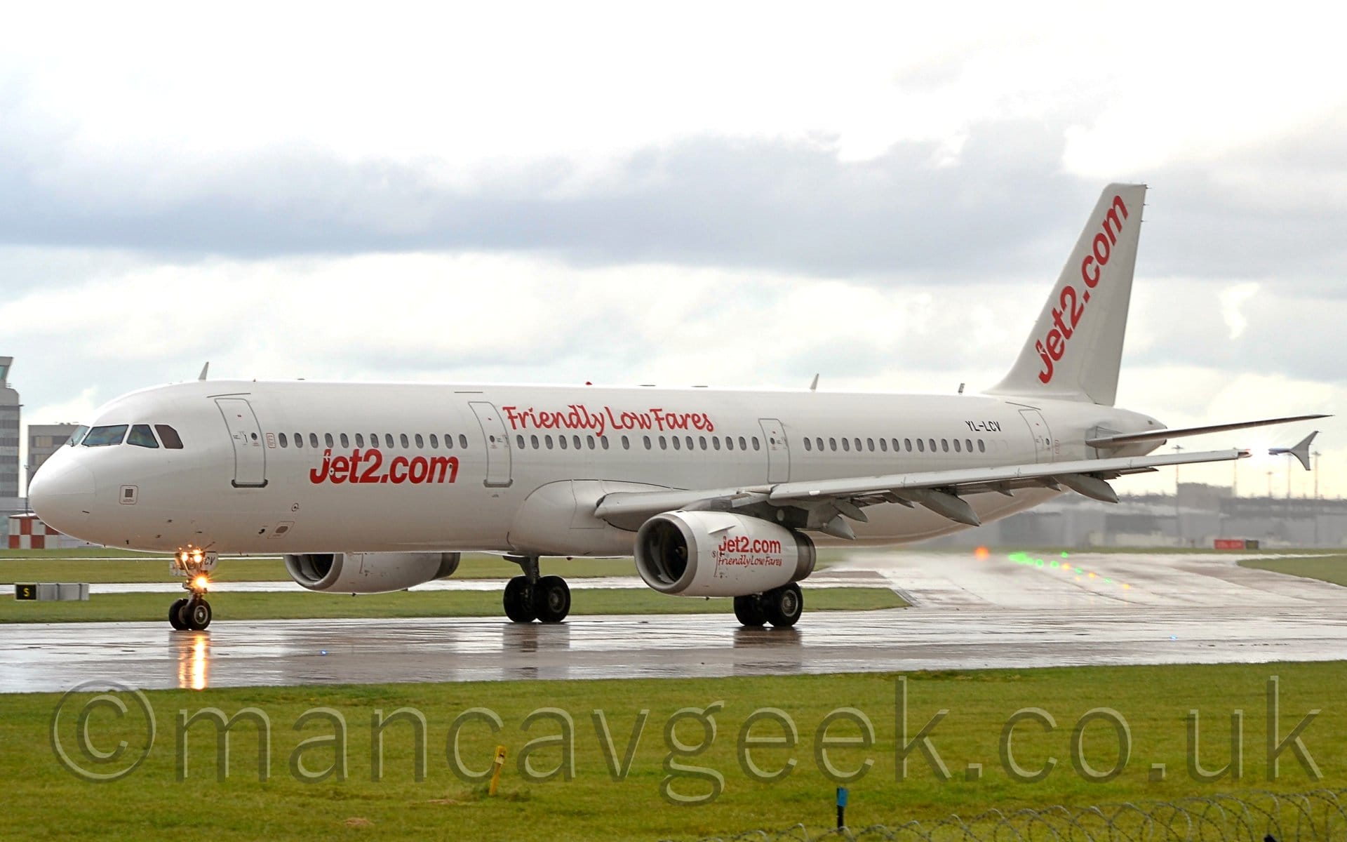 Side view of a twin engined jet airliner parked on a runway, ready for take-off. The plane is basically white, with red "Jet2.com" titles on the lower forward fuselage, red "Friendly Low Fares" text on the upper middle fuselage, over the wings, and both sets of text on the white engine pods under the wings. There are also diagonal red "Jet2.com" title on the white tail. Several lights on the nose-wheel leg are reflected on the damp runway. Grass fills the foreground, with more grass visible under the plane in the background, leading up to large terminal buildings in the distance on both sides of the frame. Bright but cloudy sky completes the image.