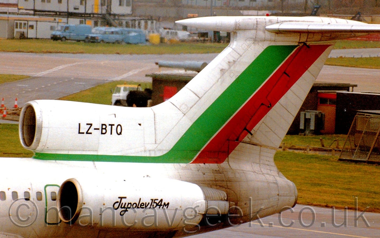 The rear fuselage of a 3 engined jet airliner, with the engines mounted on the top and sides of the rear fuselage, parked facing to the left. The plane is mostly white, with green stripe running along the top of the rear fuselage under the centre engine, sweeping diagonally up into the white tail, where it is joined by a diagonal red stripe. The black registration "LZ-BTQ" is on the centre engine intake, while the side mounted engine has black text "Tupolev 154M". A dark brown brisk building in the background is partially hidden by the tail, with a taxiway beyond that, and blue airport vans parked in front of some grey pre-fabricated buildings in the distance.