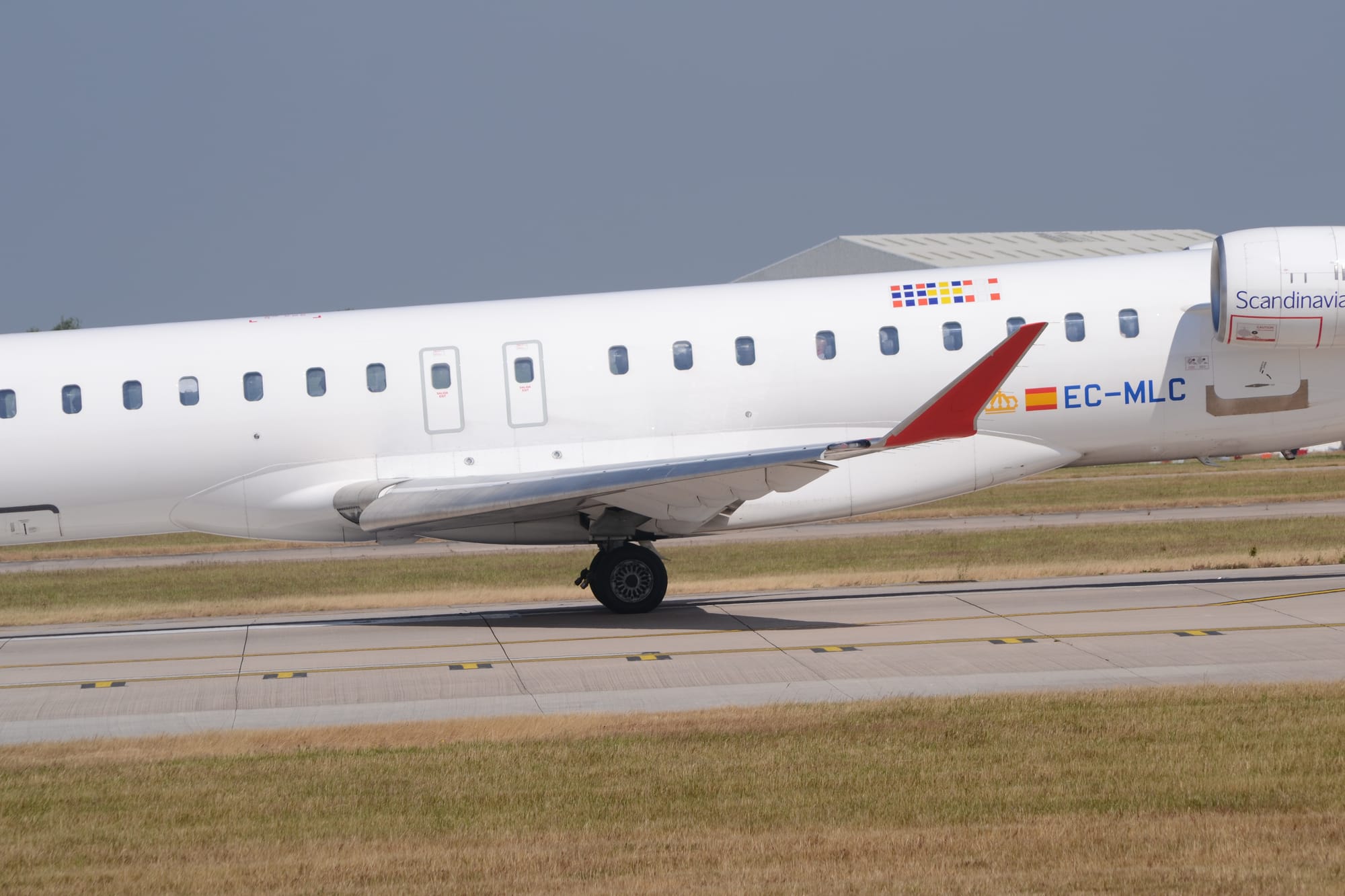 Close-up of the red up-turned wingtip of a twin engined jet airliner taxiing from right to left. The plane is almost entirely white, with a stylised yellow crown and a small Spanish flag next to the blue registration "EC-MLC"  on the lower rear fuselage, between the rear of the wing root and the white engine pods mounted on the upper rear fuselage. There are blue "Scandinavian" titles on the sides of the engine pods. Grass lines the sides of the grey runway, with the roof of a grey hangar visible over the top of the fuselage in the background, under blue-grey sky.