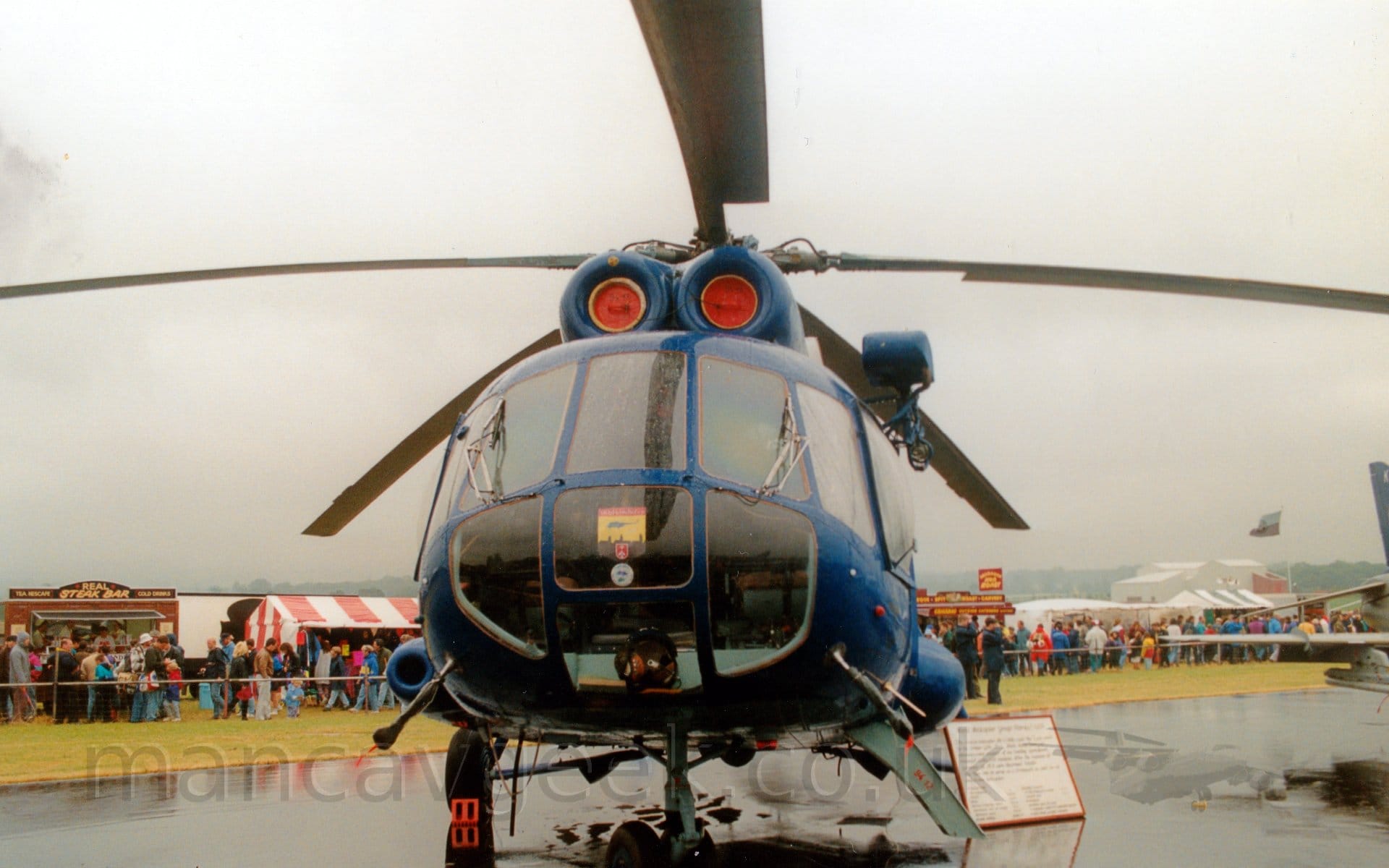 Head-on view of a dark blue helicopter with a heavily glazed nose parked on a saturated black taxiway, facing slightly left of the camera.