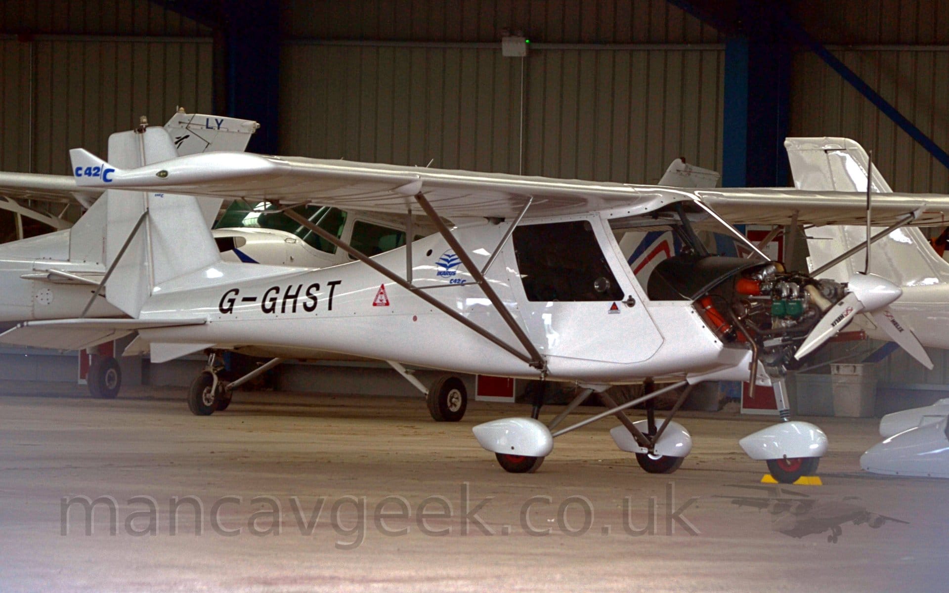 Side view of a white, high-winged, single engined light aircraft parked in a hangar facing to the right, with it's engine cowling in the nose removed, exposing it's engine.