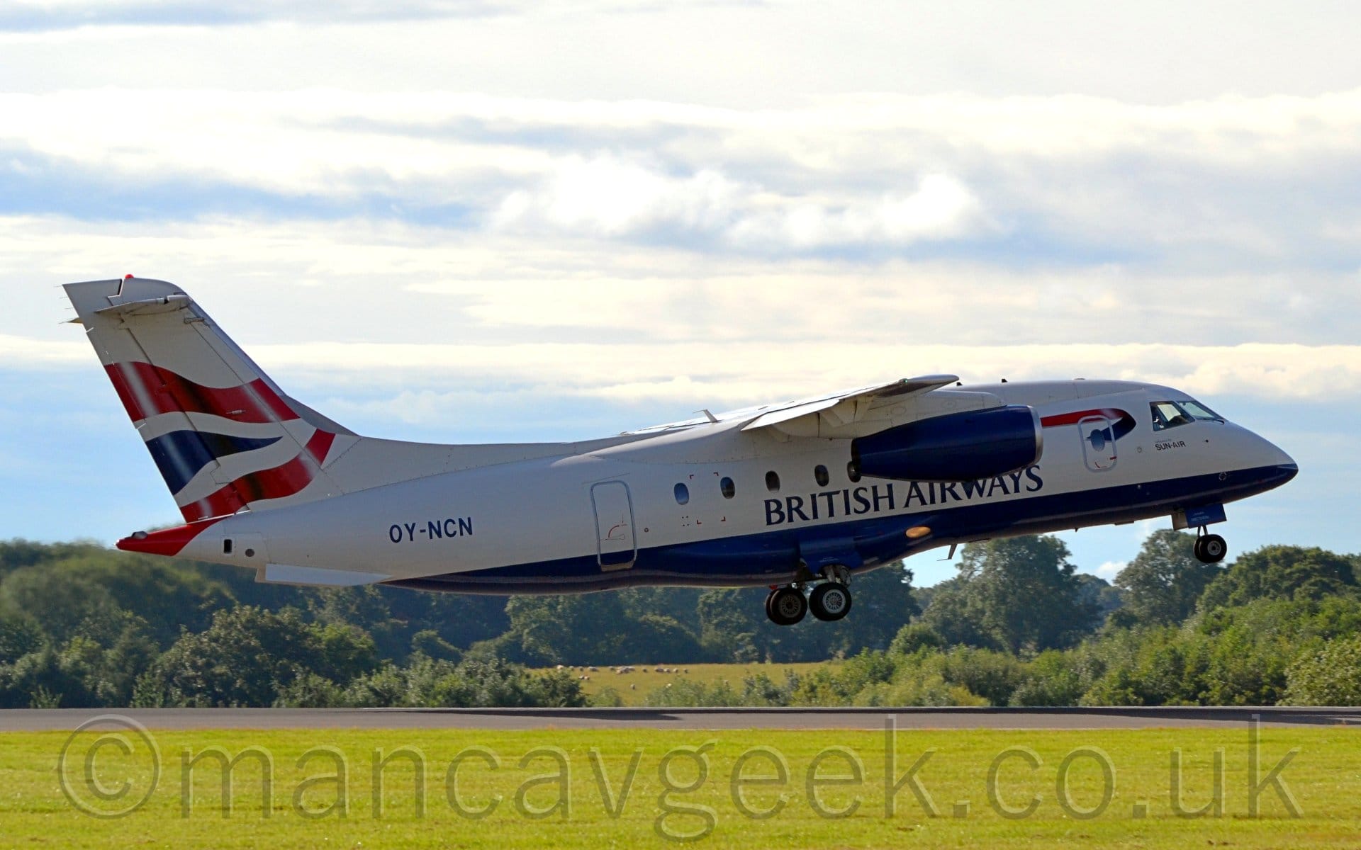 Side view of a high-winged, twin jet engined airliner flying from left to right at a very low altitude, just a couple of metres above the runway.