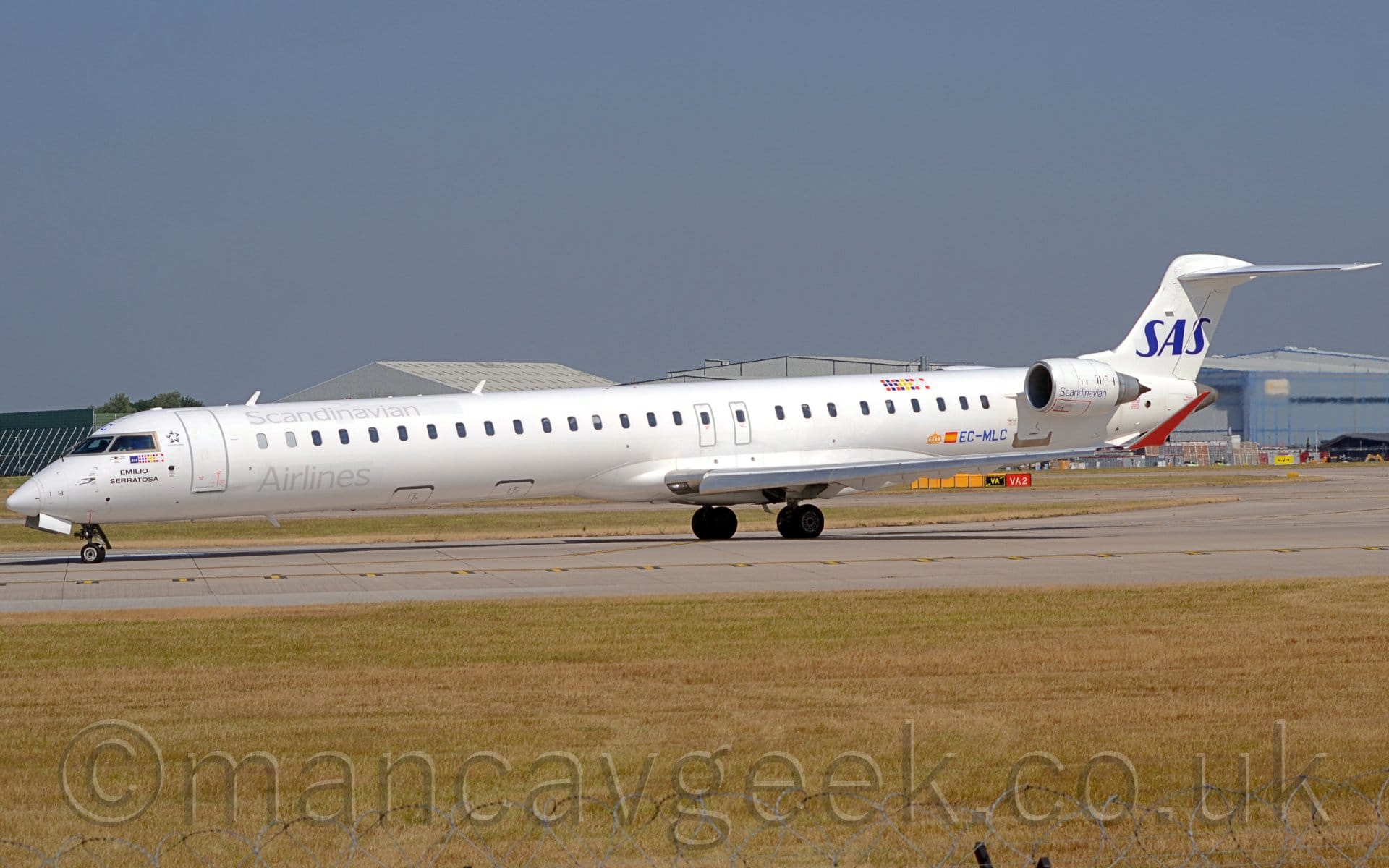 Side view of a long, thin white twin engined jet airliner taxiing from right to left as it turns on to a grey runway.
