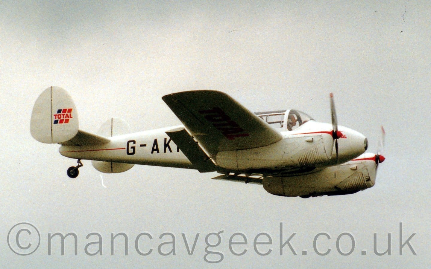 Side view of a twin engined light aircraft flying from left to right at a low altitude, with flaps extended from behind the wings.
