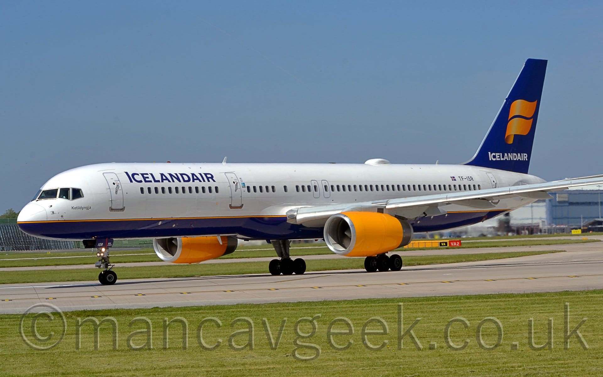 Side view of a twin engined, white and blue, jet airliner taxiing from right to left along a grey concrete taxiway