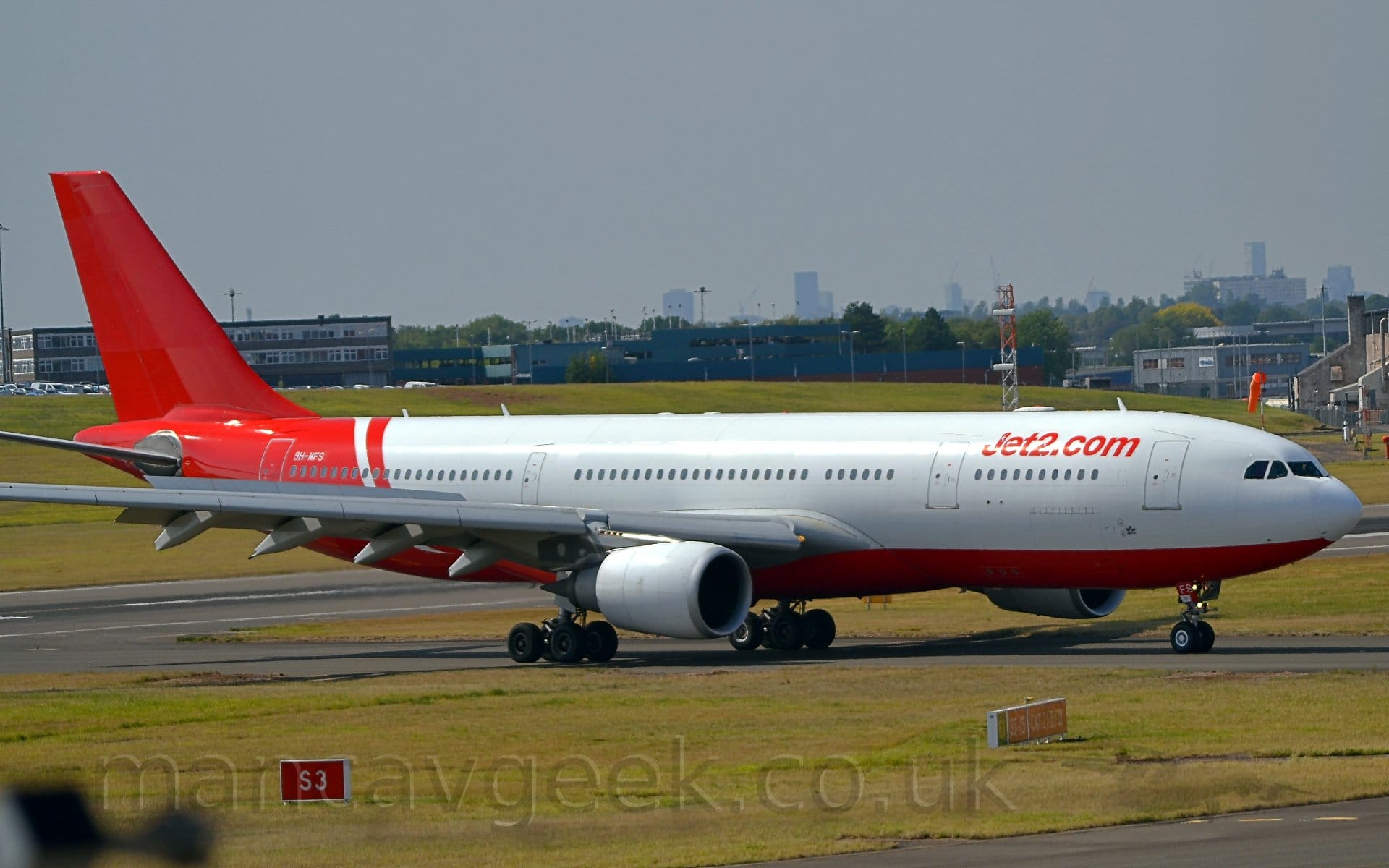 Side view of a twin engined jet airliner taxiing from left to right along a grey taxiway lined by grass. The plane is largely white, with red belly, rear fuselage, and tail.
