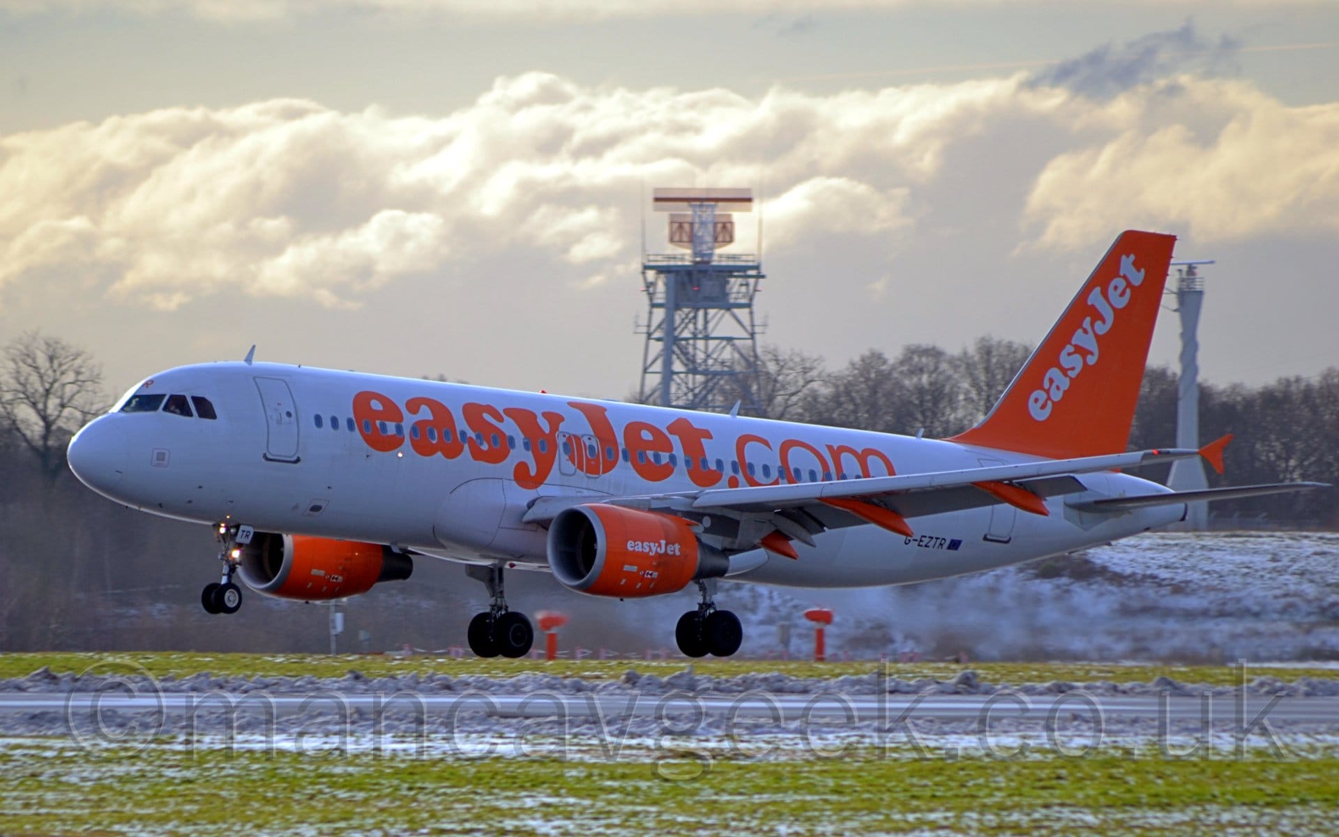 Heavily backlit side view of a twin engined jet airliner flying from right to left at a very low altitude, just a few metres above a black runway.
