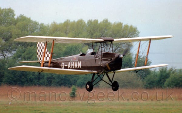 Side view of a brown, single-engined biplane flying from left to right at a very low altitude.