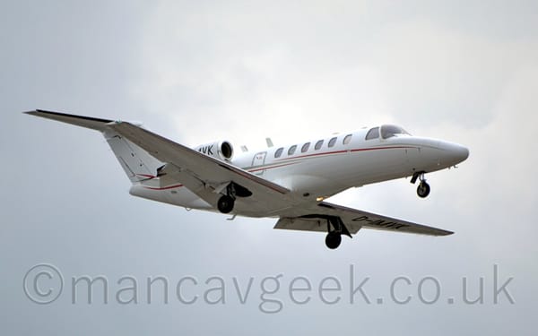 Side view of a white, twin engined BizJet with the engines mounted on the sides of the rear fuselage, flying from left to right at a low altitude