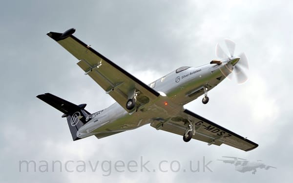 Low aside view of a silvery-grey, single engined BizProp flying from left to right at a very low altitude