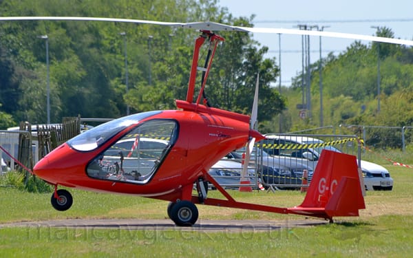 Side view of a bright red, 2 seat autogyro, parked facing to the left.
