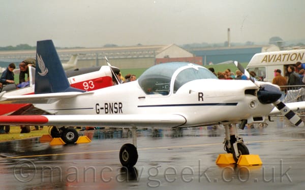 Side view of a white, single engined light aircraft with an over-sized bubble canopy over the 2 seat cockpit, parked facing to the right.
