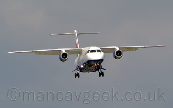 Front view of a high-winged, twin engined jet airliner flying almost head-on towards the right of the camera at a very low altitude