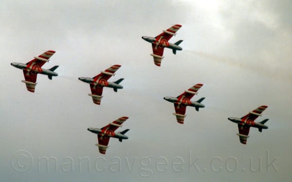 View from below of 6 single engined fighter jets flying in close formation from right to left at a low altitude.