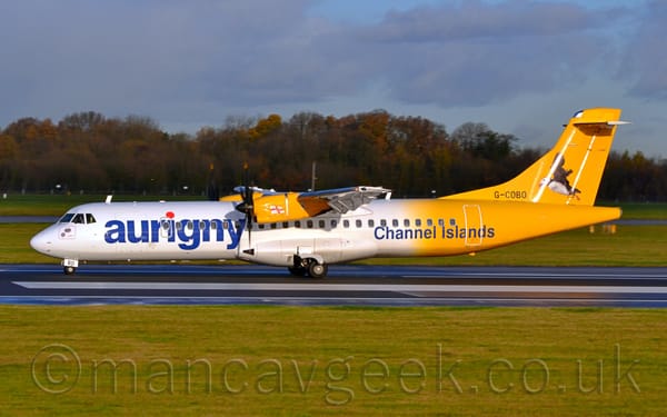 Side view of a high-winged, twin propellor-engined, white and yellow airliner moving from right to left along a black runway.