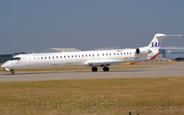 Side view of a long, thin white twin engined jet airliner taxiing from right to left as it turns on to a grey runway.