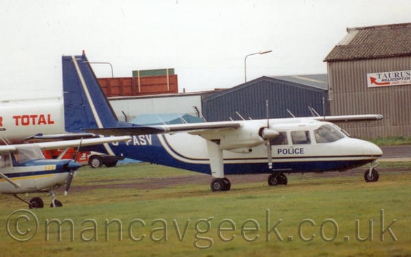 Side view of a high-winged, twin propellor engined light transport plane parked on grass facing to the right.