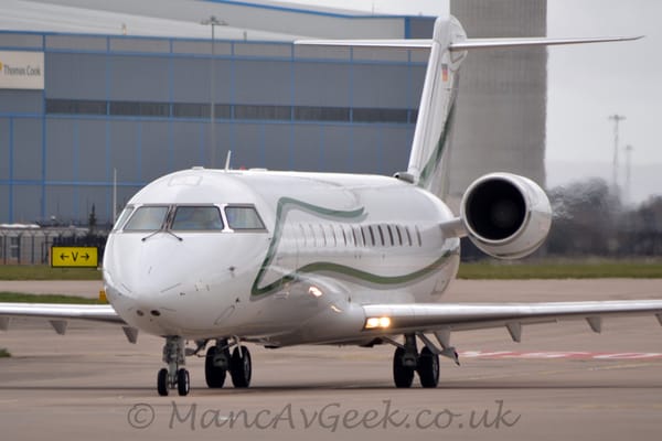 Front view of a white, twin engined BizJet taxiing from right to left towards the camera on a wide grey taxiway.