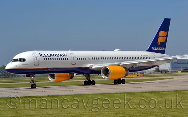 Side view of a twin engined, white and blue, jet airliner taxiing from right to left along a grey concrete taxiway