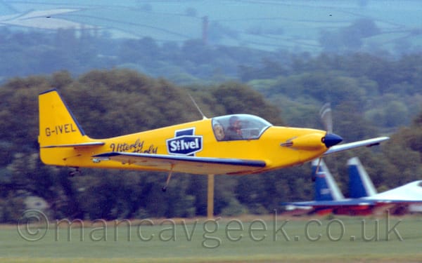 Slightly grainy side view of a bright yellow, single-engined light aircraft flying from left to right at a very low altitude.