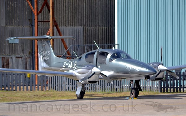 Side view of a rather sleek, twin engined light aircraft parked on grey tarmac facing to the right.