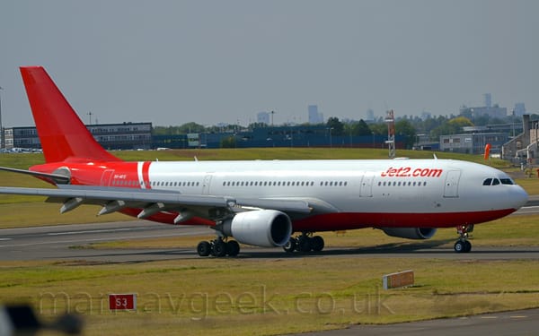 Side view of a twin engined jet airliner taxiing from left to right along a grey taxiway lined by grass. The plane is largely white, with red belly, rear fuselage, and tail.