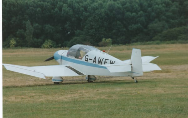 Rear side view of a single-engined, white light aircraft parked on grass facing to the left