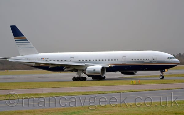 Side view of a twin engined jet airliner taxiing from left to right along a black taxiway.