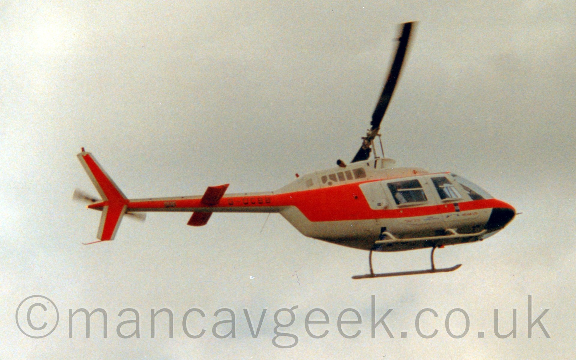 Side view of a helicopter flying from left to right at low level. It's body is mostly cream, with reddish-orange stripe running from the nose and up in to the tail boom, where it covers most of the tail. The main and tail rotors appear to be almost frozen in place. Behind, the sky is a dreary gray.