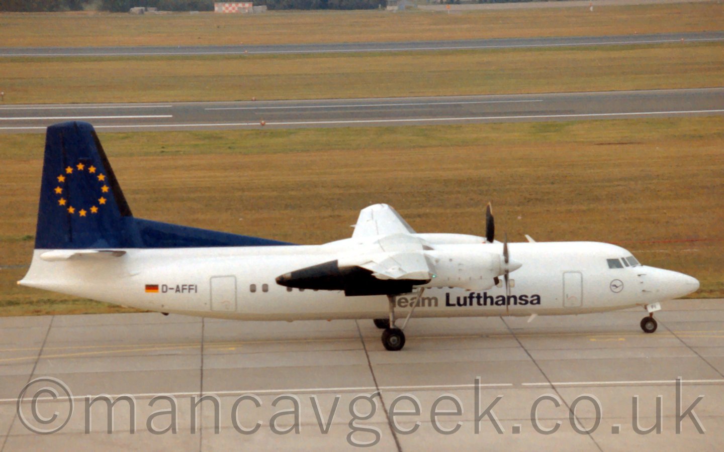 Side view of a twin propellor engined, high-winged airliner taxiing from left to right. The planes body is mostly white, with large "Team Lufthansa" titles on the lower fuselage in grey and dark blue, while the tail is dark blue with a ring of 12 5-pointed gold stars. The background is mostly grass, with a couple of strips of runway visible.