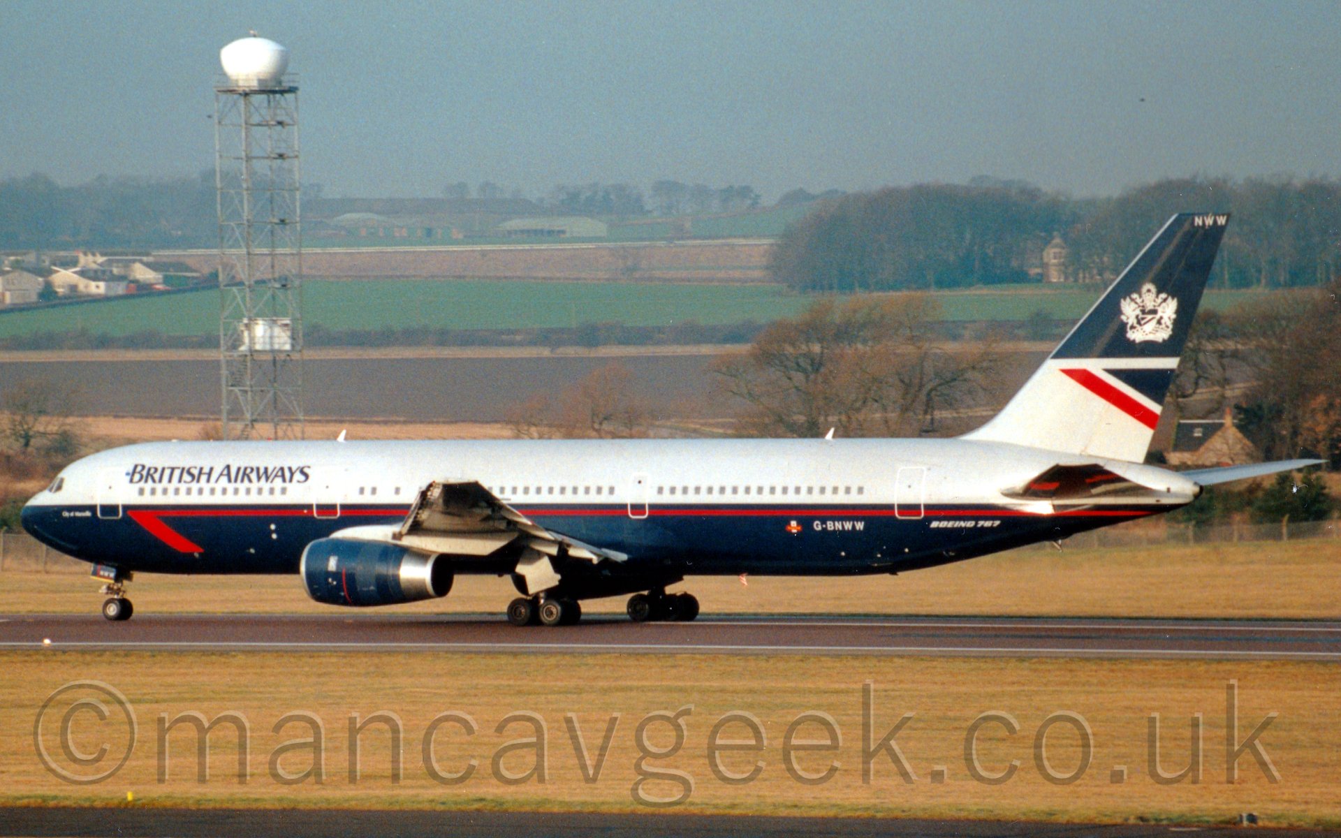 Side view of a twin engined jet airliner running along a runway from right to left at high speed during it's take off run. The planes body has a dark blue belly with a thin red stripe, while the upper half is grey, with dark blue "British Airways" titles on the upper forward fuselage. The tail has a blue top, with a promiently displayed Royal Crest, and a grey lower half, with a blue, grey, and red triangle. The front of the frame is taken up with grass, while fields lead off into the distance, giving way occasionally to trees and bushes. There is a large tank on a tall metal tower just behind the planes cockpit.