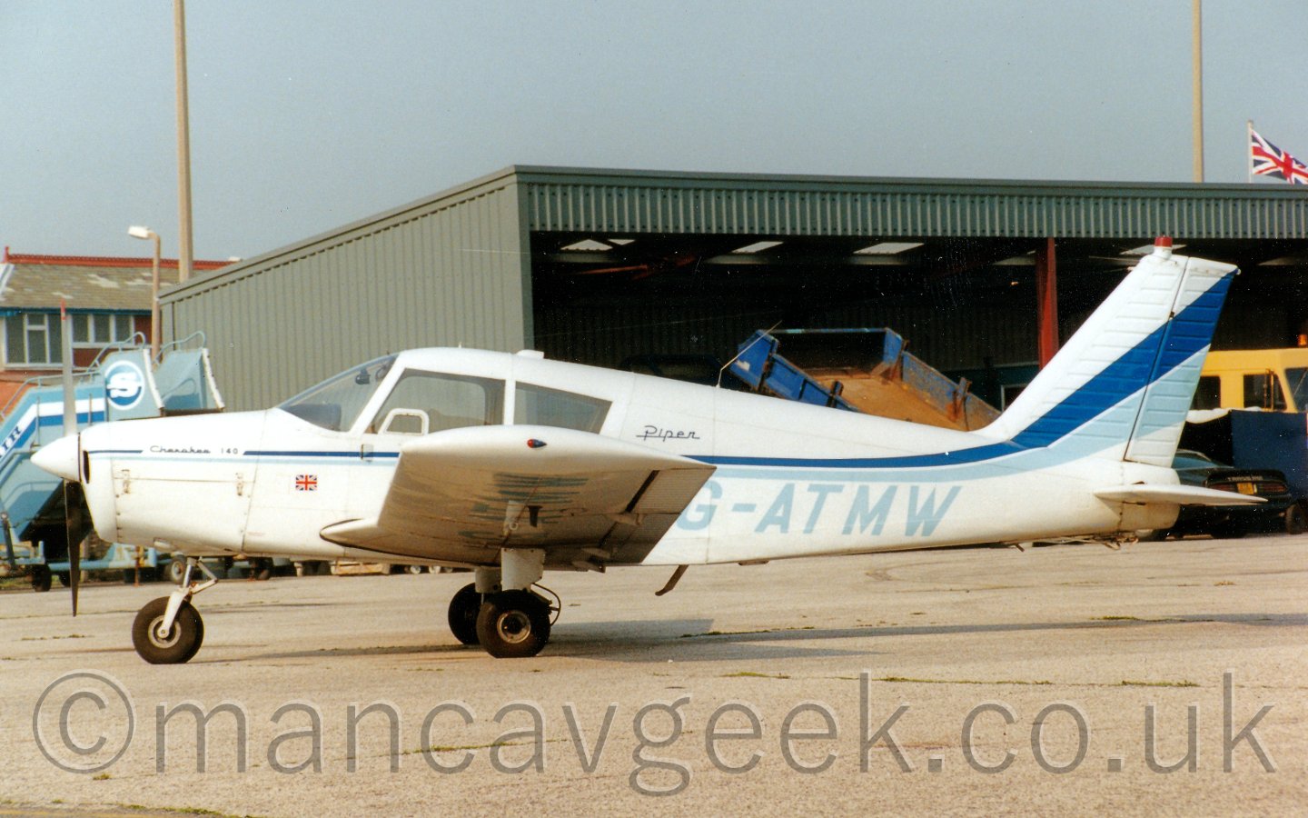 Side view of a single propellor engined light aircraft parked in front of a metal grey building containing some airport vehicles. The planes body is mostly white, with a two-tone blue strip running backwards from the nose before sweeping up into the tail, getting wider as it goes.