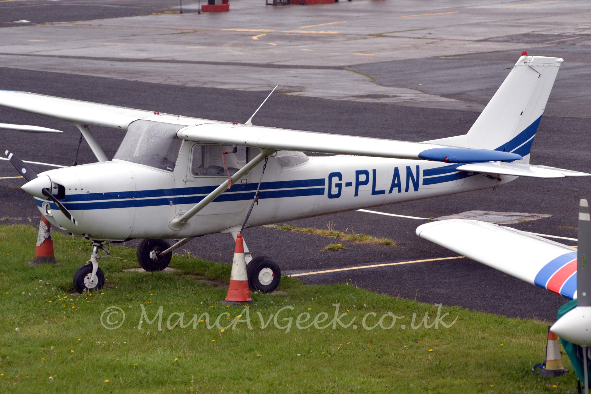 Side view of a single propellor-engined light aircraft parked on grass at the edge of a tarmaced area between a pair of orange and white traffic cones. The plane is mostly white, with a pair of blue stripes running back along the fuselage from the nose, with the registration "G-PLAN" on the rear fuselage. There is the wing and engine of another light aircraft just peeking in to view on the lower right of the frame.