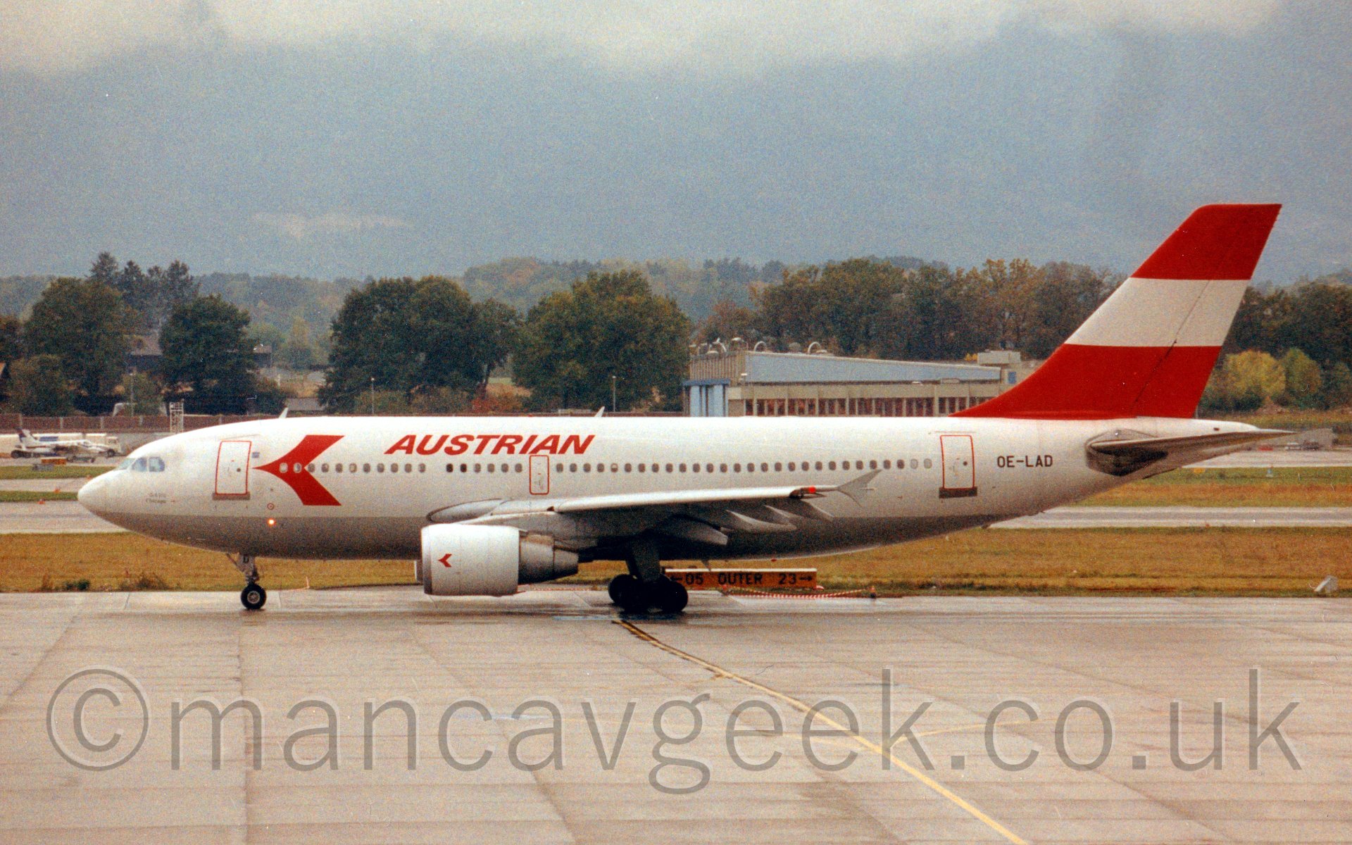 Side view of a twin engined jet airliner taxiing from right to left. The plane is mostly white, with a silver belly, and a large red arrowhead on the forward fuselage, with red "Austrian" titles immediately after. The tail is mostly red, with a thick white stripe. Behind is a view across the runway to a hangar on the far side, with some light aircraft parked in front.