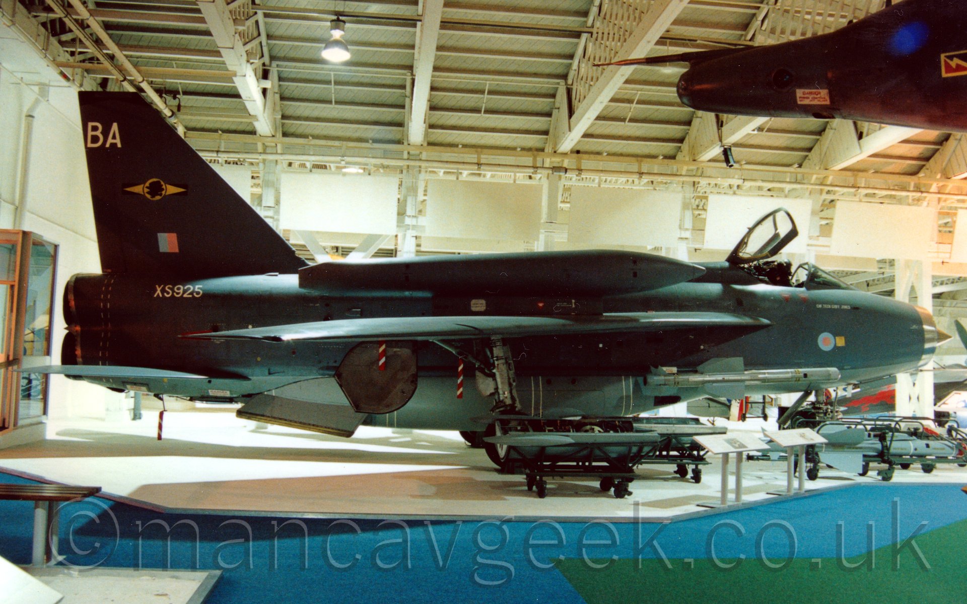 Side view of a single-seat twin engined jet fighter aircraft on display in a museum. The plane has a grey body, with a silver nose cone, with an open cockpit canopy. The plane has a large fuel tank mounted over the wing. It carries the serial "XS925" in whiute on the upper rear fuselage, has a small blue and red square flag and large letters "BA" on the tail, and a small blue and red roundel on the nose. The nosecone of another plane is just visible in the top right corner. Behind are the white walls and ceiling of the museum