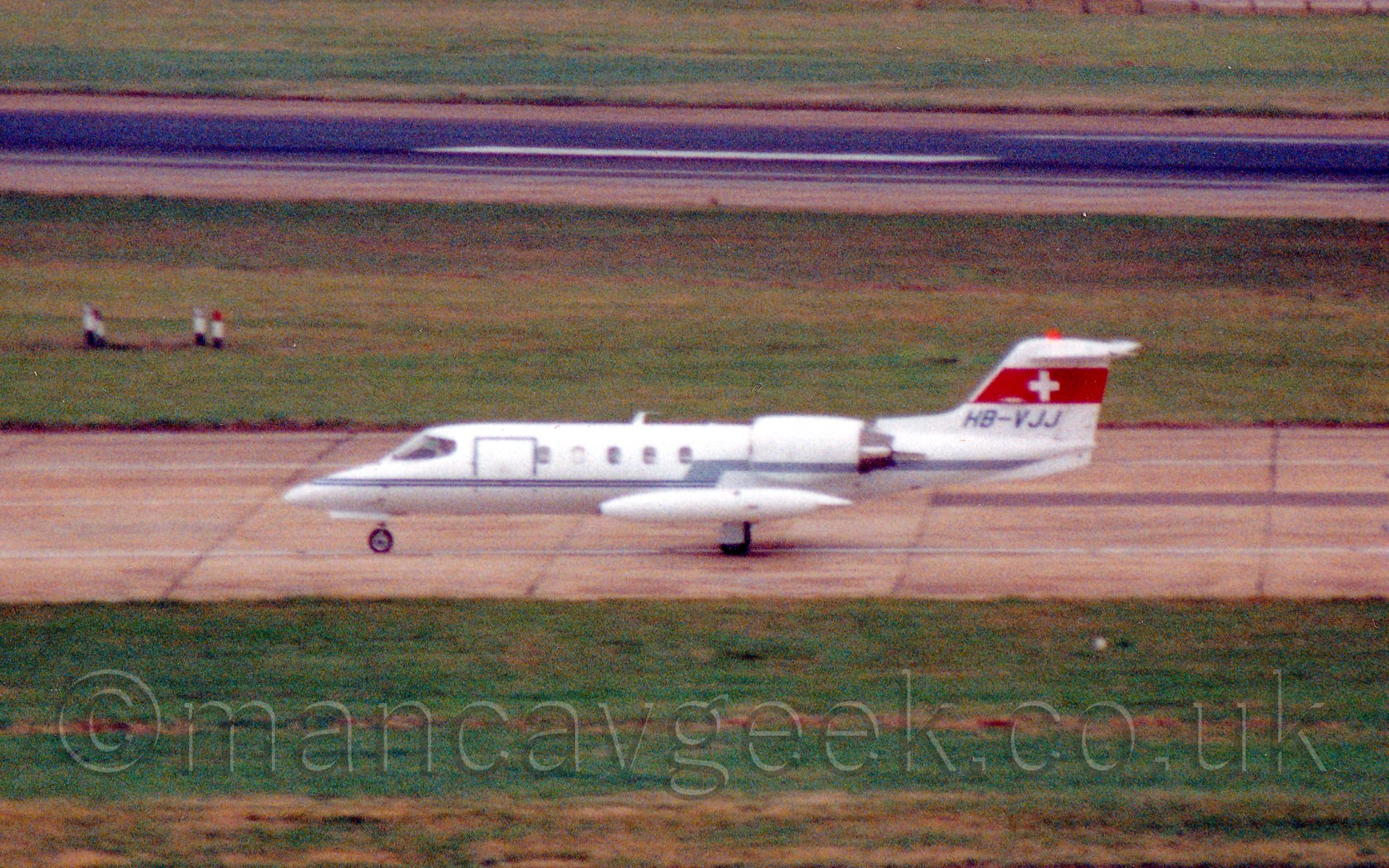 Side view of a twin engined bizjet taxiing from right to left. The planes body is mostly white with a blue line running from nose to tail, rising slightly over the wing to also cross the engines. There is a thick red stripe 2/3rds of the way up the tail with a white cross in the middle. The background