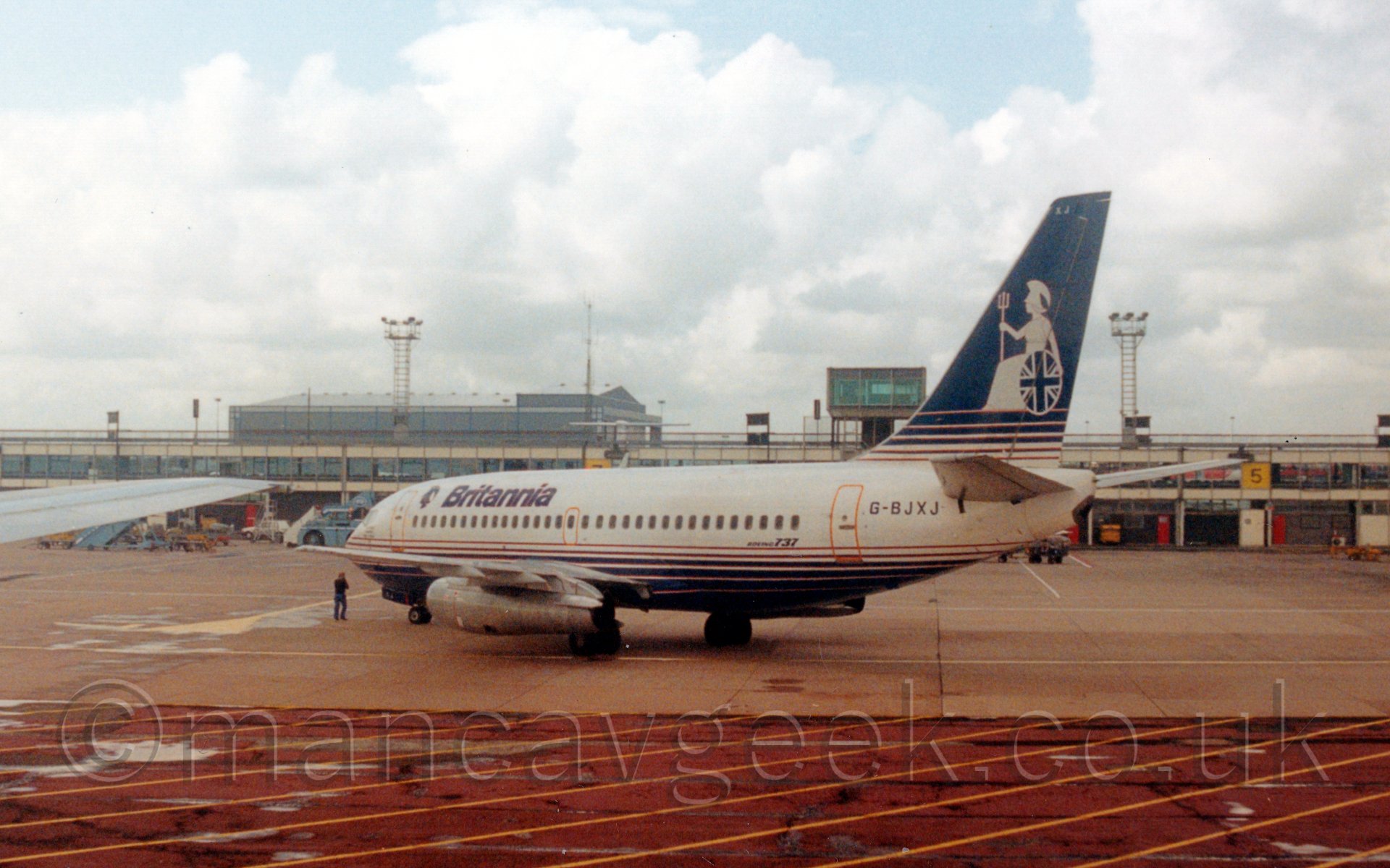 Side view of a twin engined jet airliner being pushed back from it's stand at an airport. The plane's body is mostly white, with a blue belly and blue stripes on the lower fuselage, with blue "Britannia" titles on the upper forward fuselage. The tail is mostly blue with white stripes at the bottom, with an image of a seated Britannia. In the background, one of the airport piers stretches from one side of the frame to the other, with a large grey hangar on the left, while above, fluffy grey clouds are giving way to blue.