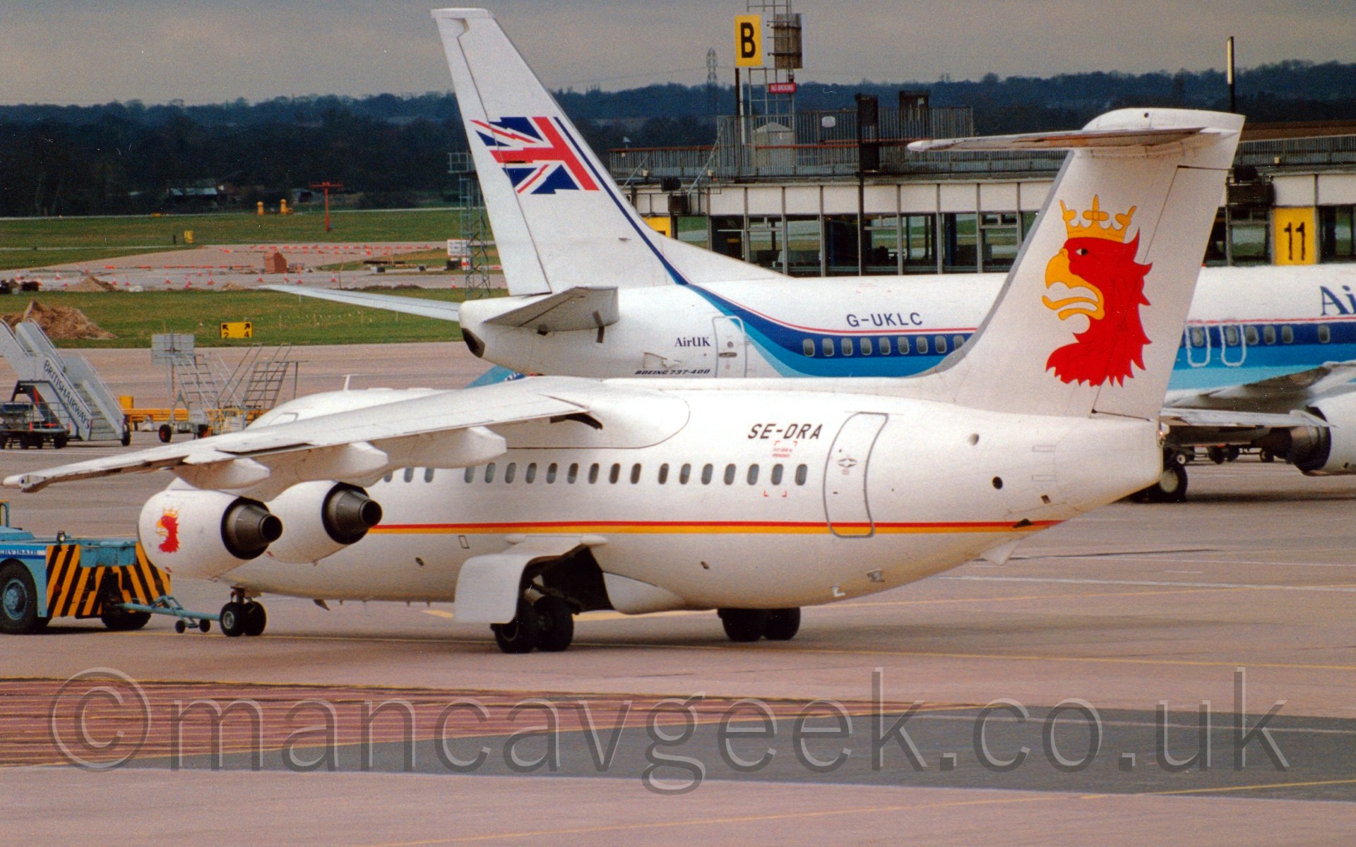 Side view of a high-winged 4 engined jet airliner being puhed off it's stand, facing to the left of frame. The plane is white, with a thin red and yellow stripe running along the fuselage, and an anthopomorphised painting of the head of a red bird with a golden beak, wearing a gold crown. The birds beak is open, with it's tonguewaving. The planes registration "SE-DRA" is prominently displayed on the upper rear fuselage, just forward of the aft door. There is a low-slung light blue vehicle with diagonal yellow and black stripes on its rear attached by a blue bar to the nose of the plane, slightly out of frame on the left. Behind is a slightly larger white twin engined jet airliner with a three-tone blue stripe running along the fuselage, swooping up in to the tail where it becomes a flagpole for a waving half British flag,behind which is a terminal building. In the background, large grassed areas give way to trees in the distance, under a grey sky.