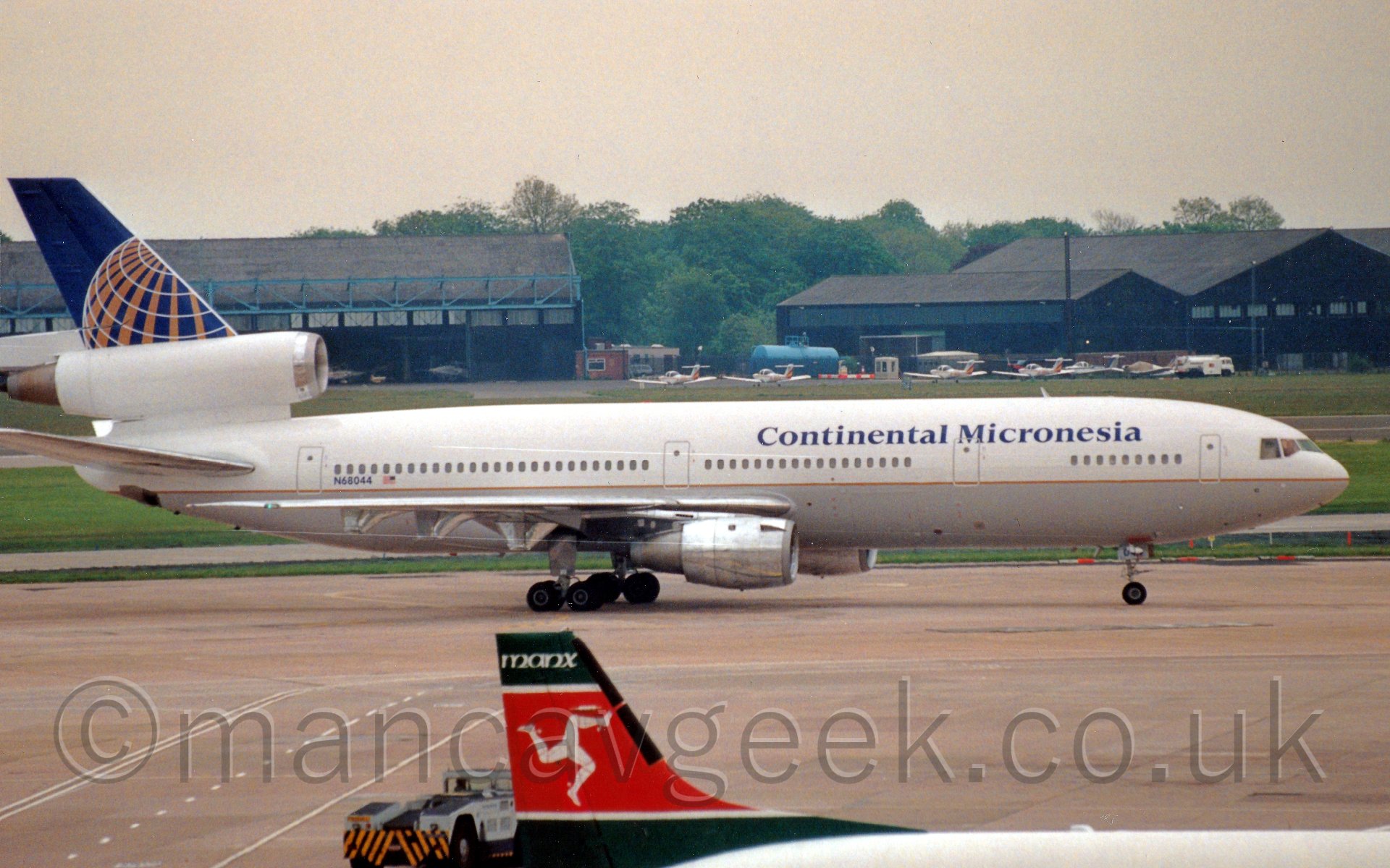 Side view of a three engined jet airliner taxiing from left to right. The plane has a light grey belly and white upper half, seperated by a thin gold stripe. There are dark blue "Continental Micronesia" titles on the upper forward fuselage. The tail has a white lower section, up to the top of the centre engine cowling, then the upper section is a dark blue, overlaid with a stylised wireframe globe in white and gold. In the foreground is the tail of a smaller plane, mostly red, with green stripes at the top and bottom. The red section has 3 legs, arranged like the spokes of a wheel, joined at the hip and looking as if they are running/rolling. In the background are a handful of black hangars, with some light aircraft parked in front of them, with trees behind. under a yellow-tinged grey sky