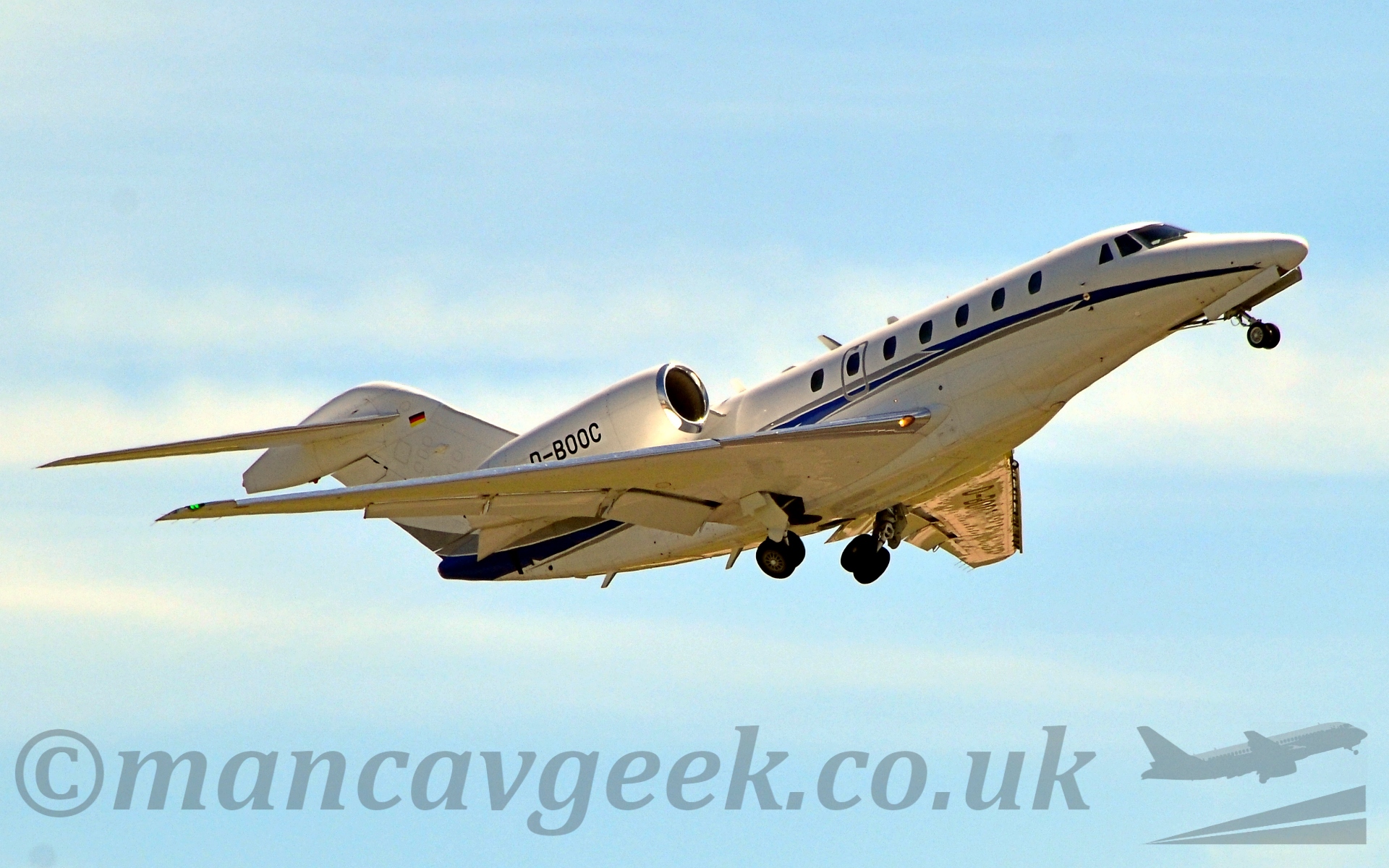 Low side view of a twin engined bizjet flying from left to right at a very low altitude, withflaps deplyed, landing gear being tucked away, and nose raised, suggesting it has just taken off. The plane is mostly white, with a blue and gresy stripe zig-zagging it's way along the fuselage and up into the base of the tail. The registration "D-Booc" is prominent of the engine cowlings, and there is a small German flag (black, red, and yellow horizontal bands) at the top of the tail. Behind, the sky is a light blue, with white clouds in the far distance.